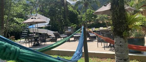 Una hamaca, sillas de descanso y sombrillas en un entorno de resort, con palmeras y una piscina.
