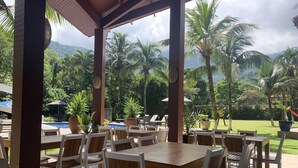 A covered outdoor dining area with wooden tables and chairs, a pool, and lush greenery in the background. - Delta Maresias Club by SmartSharing (São Sebastião)
