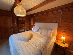 A wooden cabin bedroom with a large bed, a hanging light fixture, and a bedside table with a lamp.