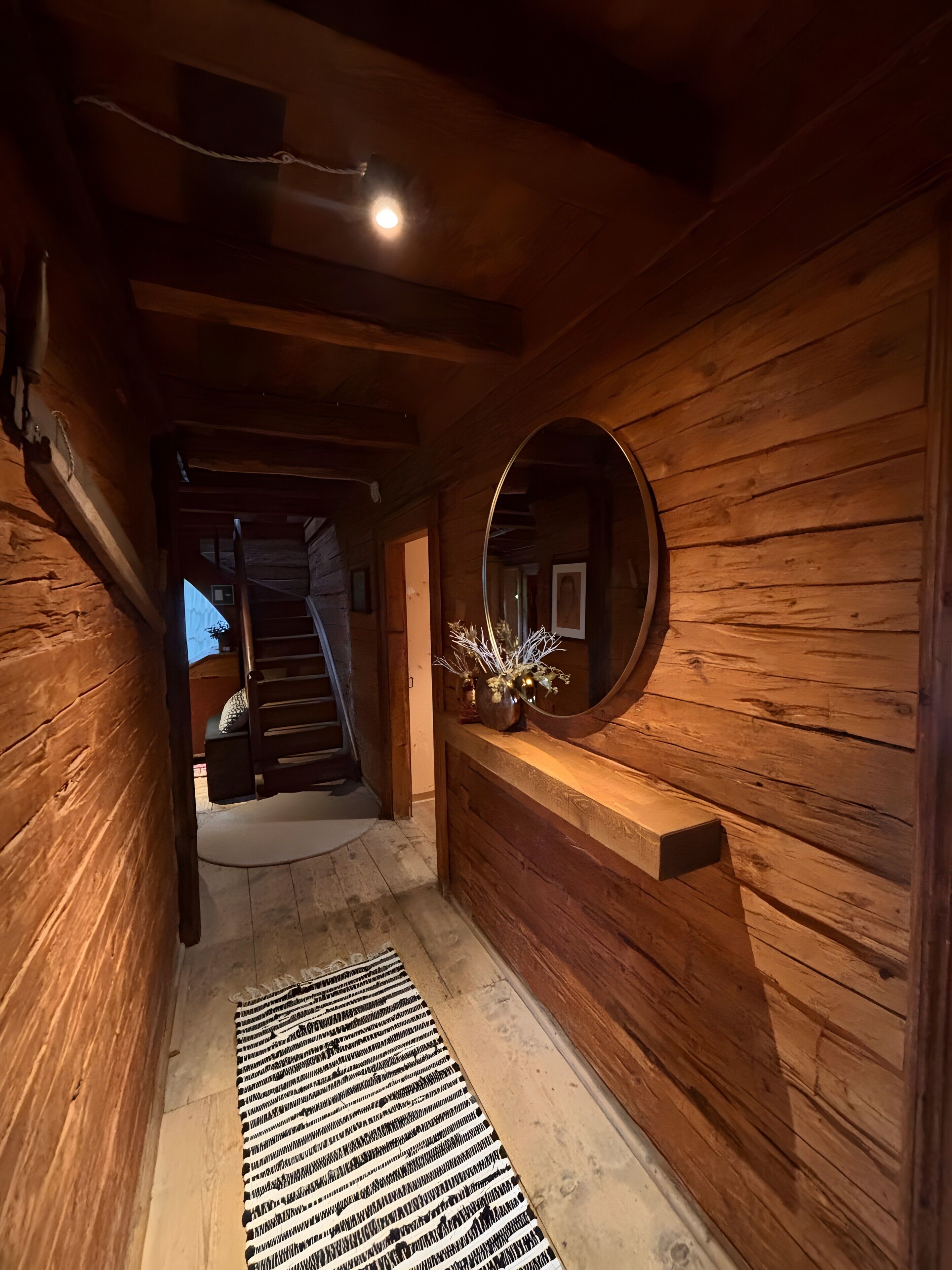 A wooden interior hallway with a mirror, a rug, and a staircase leading to another room.