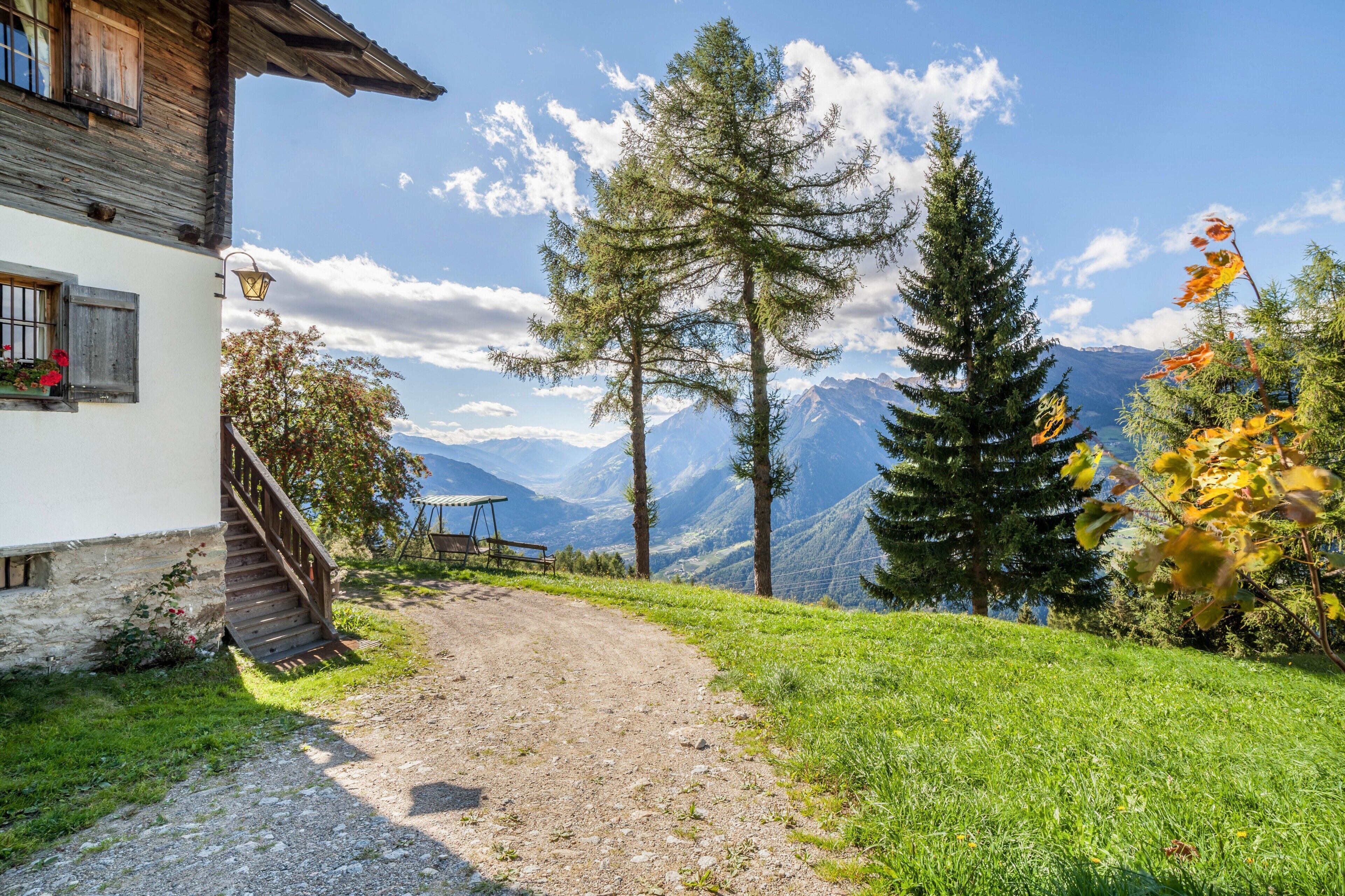 Eine Berghütte mit einem Feldweg, der zu einer Bank und einem Tisch führt, umgeben von Bäumen und Blick auf entfernte Berge.