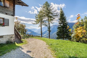 A mountain cabin with a dirt path leading to a bench and a table, surrounded by trees and a view of distant mountains.