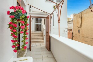 Un estrecho pasillo exterior con una escalera que lleva a una puerta, un arreglo floral colgante y una pared blanca.