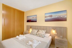 Una cama bien hecha con toallas, cabecera de madera, dos mesitas de noche con lámparas y dos cuadros de paisajes en la pared.