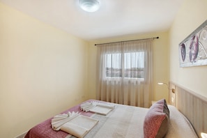A hotel room with a bed, a window with sheer curtains, a wall-mounted artwork, and a ceiling light.