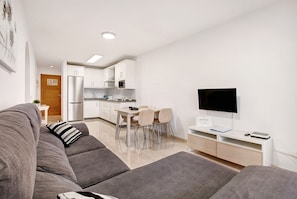 A modern living room with a sofa, a dining area with chairs, a kitchen with white cabinets, and a flat-screen TV.