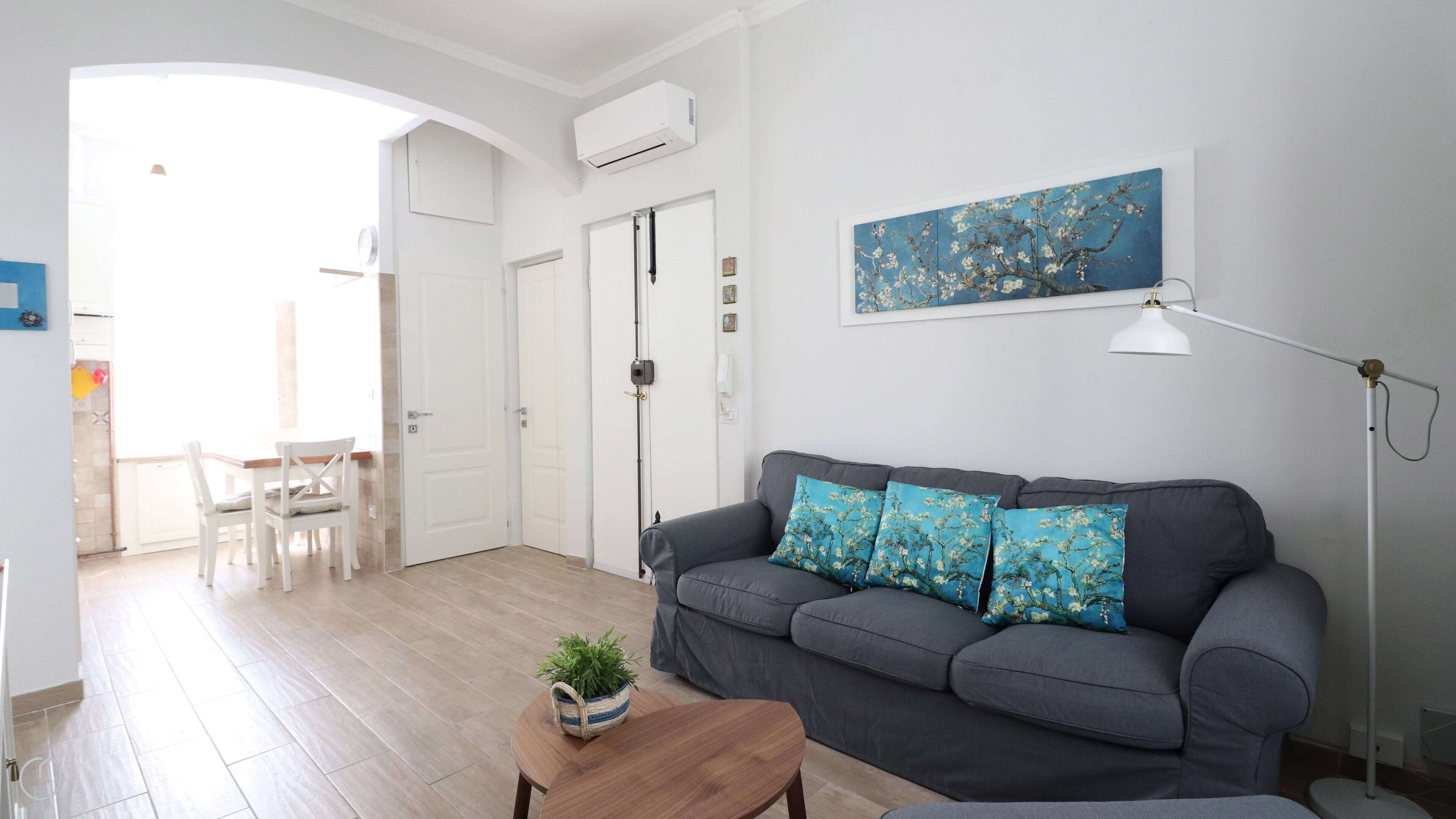 A modern living room with a grey sofa, two blue floral cushions, a wooden coffee table with a plant, a white floor lamp, and a painting on the wall.