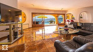 A spacious living room with a large flat-screen TV, a glass coffee table, and a leather sofa. The room features a marble floor and large windows offering a view of the sea. - Villa DIANA by Abahana Villas (Teulada)