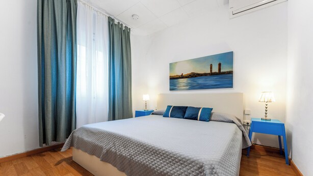 A neatly arranged hotel room with a large bed, two bedside tables with lamps, a painting on the wall, and a view of the cityscape through the window. - Tranquilo. Cerca de la Playa. Bien Comunicado (Sant Adrià de Besòs)