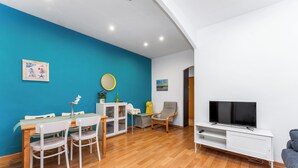 A modern living room with a blue accent wall, wooden flooring, a dining area with white chairs, a TV on a white stand, and a grey sofa. - Tranquilo. Cerca de la Playa. Bien Comunicado (Sant Adrià de Besòs)