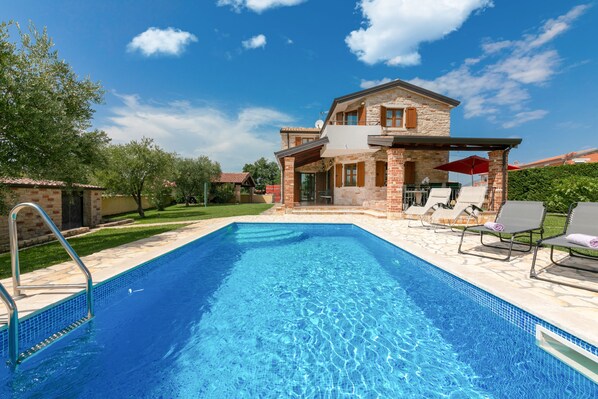 A swimming pool with a sun terrace, lounge chairs, and a stone house with a balcony.