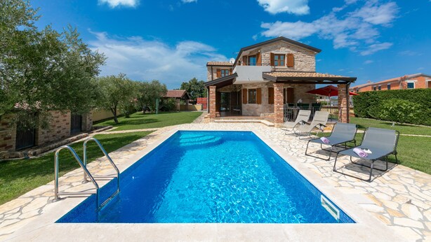 Une piscine rectangulaire bordée d’une terrasse en pierre, entourée de chaises longues et d’une maison en pierre dotée d’un patio.