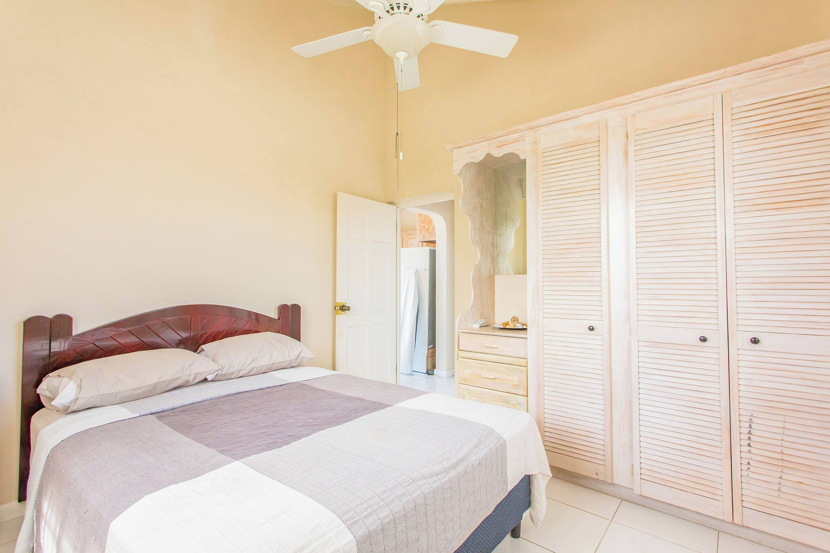 A bedroom with a bed, a ceiling fan, a wardrobe, and a door leading to another room.