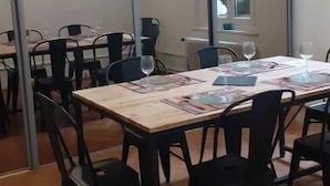 Uma sala de jantar com uma mesa grande posta para quatro pessoas, rodeada por cadeiras. Há uma porta de vidro deslizante que dá para outro espaço, e uma luminária pendente ilumina a mesa.