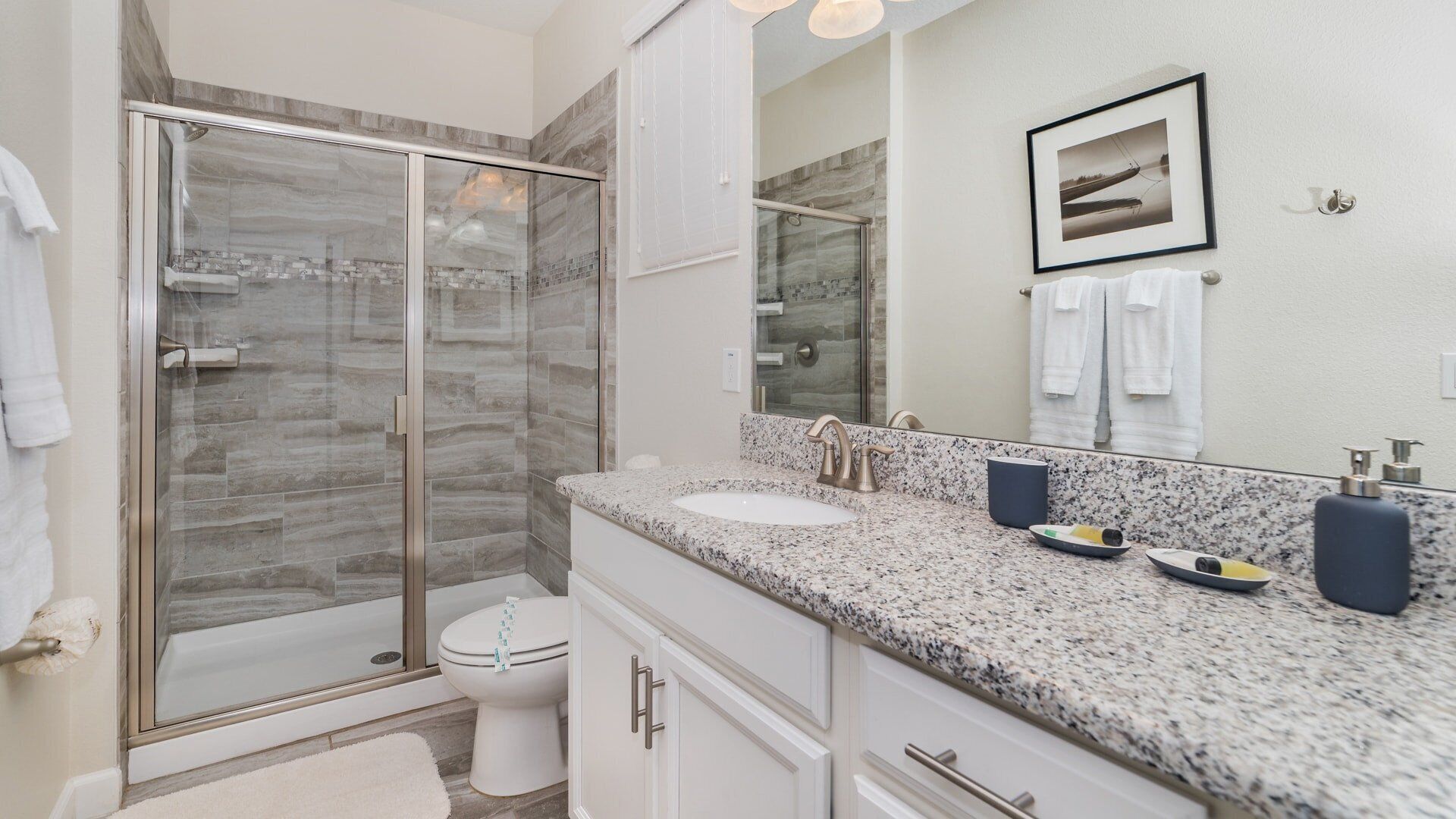 A modern bathroom with a glass-enclosed shower, a white toilet, a granite countertop, and a large mirror.