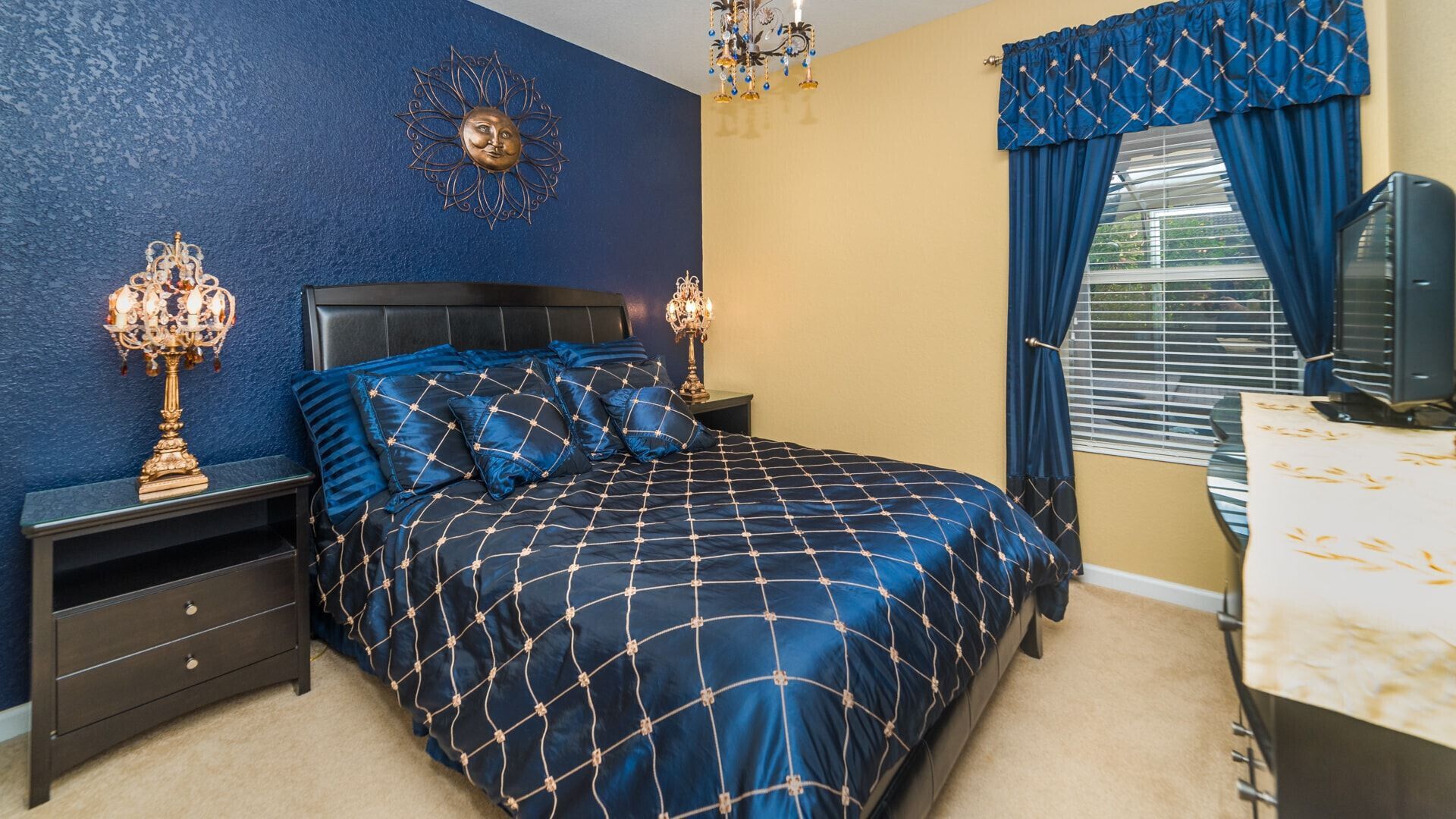 Un dormitorio con cama, mesitas de noche, cómoda, candelabro y ventana con cortinas.