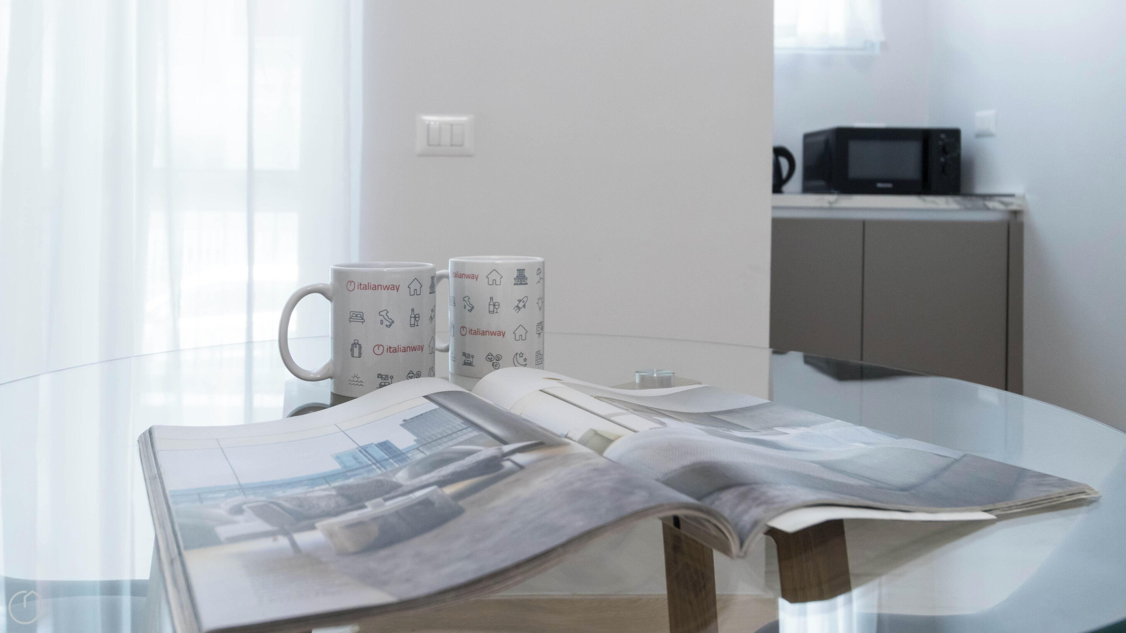 A glass table with newspapers, two Italianway branded mugs, and a microwave in the background.