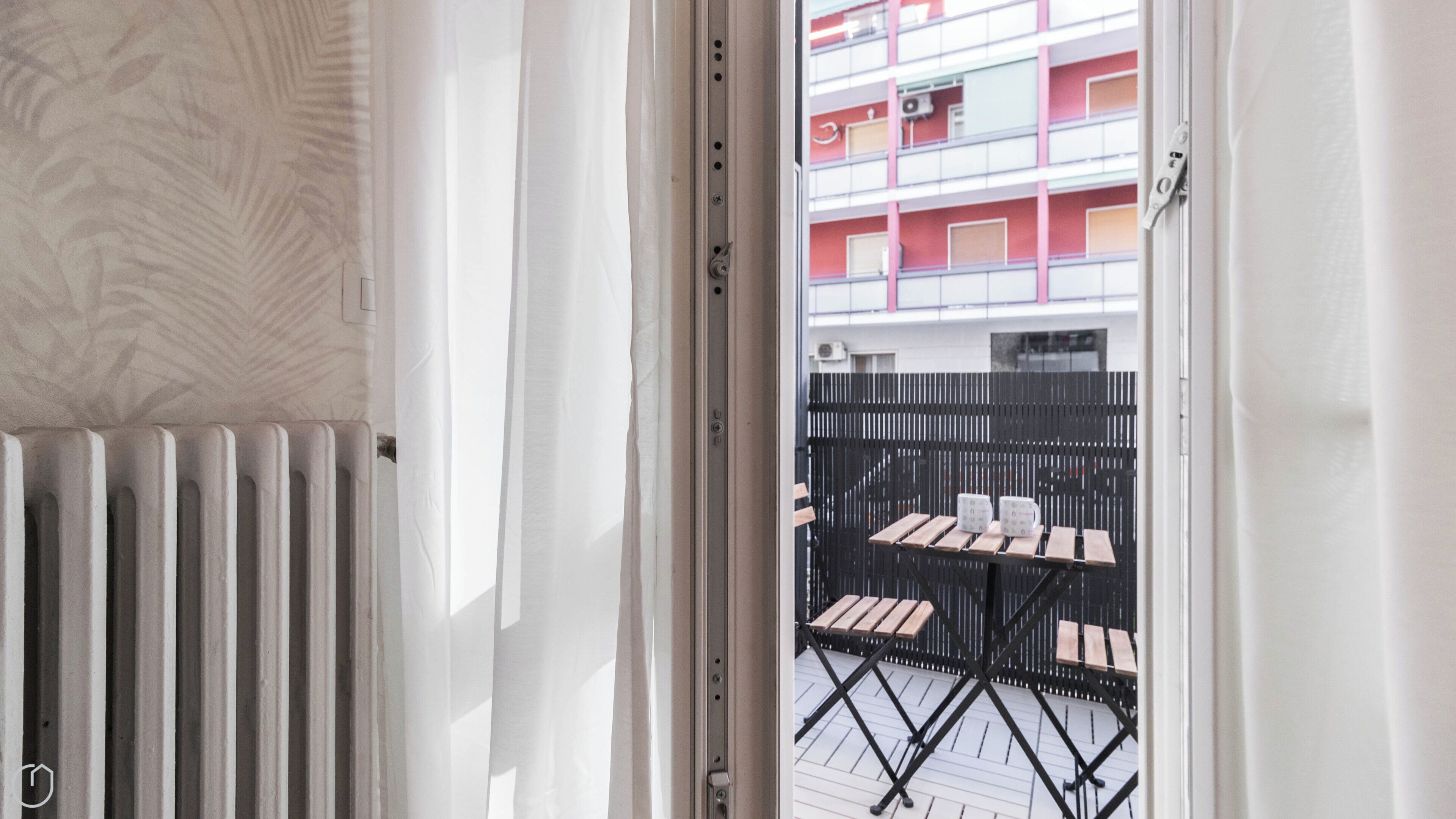 A view through a window with a radiator, a black metal fence, and a table with chairs outside.