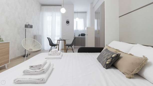 A modern hotel room with a large bed, a desk, and a chair. There is a kitchenette in the background. - Gioberti 11 (Sesto San Giovanni)