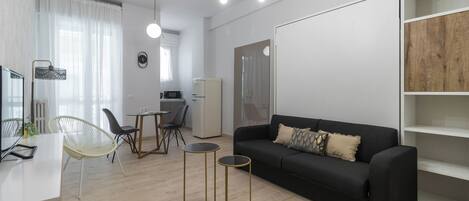 A modern living room with a black sofa, two small side tables, a dining area with a table and chairs, a kitchen area with a refrigerator, and a large window with sheer curtains.
