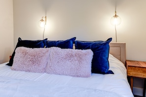 A neatly made bed with a white duvet, pink fluffy pillows, and dark blue velvet pillows. There are two wall-mounted lights on either side of the bed.