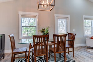 A dining area with a glass table surrounded by wooden chairs, a chandelier, and a view of trees through the windows. - Cozy and Clean, Pet and Family-Friendly Gem; Minutes to Asheville (Weaverville)