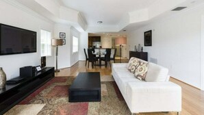 Un salon moderne avec un téléviseur à écran plat, un canapé blanc, une table basse et un coin repas à l’arrière-plan.