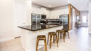 A modern kitchen with white cabinets, stainless steel appliances, a black countertop, and gold bar stools. - Last Minute Disney! Private Pool, Hot Tub, Game Room & Themed Bedrooms. Book Now! #31744 (Orlando)