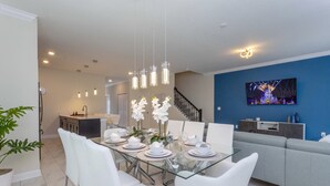 A modern dining area with a glass table set for four, white chairs, and a chandelier. A staircase leads to another room, and a TV is mounted on the blue wall. - Last Minute Disney Getaway! Private Pool & Themed Bedrooms. Book Now! #40229 (Orlando)