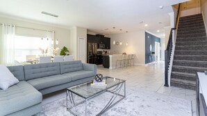 A modern living room with a grey sofa, a glass coffee table, and a kitchen in the background. - Last Minute Disney Getaway! Private Pool & Themed Bedrooms. Book Now! #40229 (Orlando)