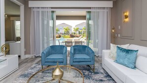 Un salon moderne avec un canapé blanc, deux fauteuils bleus, une table basse en verre et une vue sur une piscine et des bâtiments en arrière-plan.