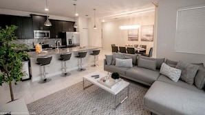 Een moderne keuken met donkere kasten, een centraal eiland met barkrukken, een eethoek met tafel en stoelen, en een woonkamer met een grijze bank en salontafel.