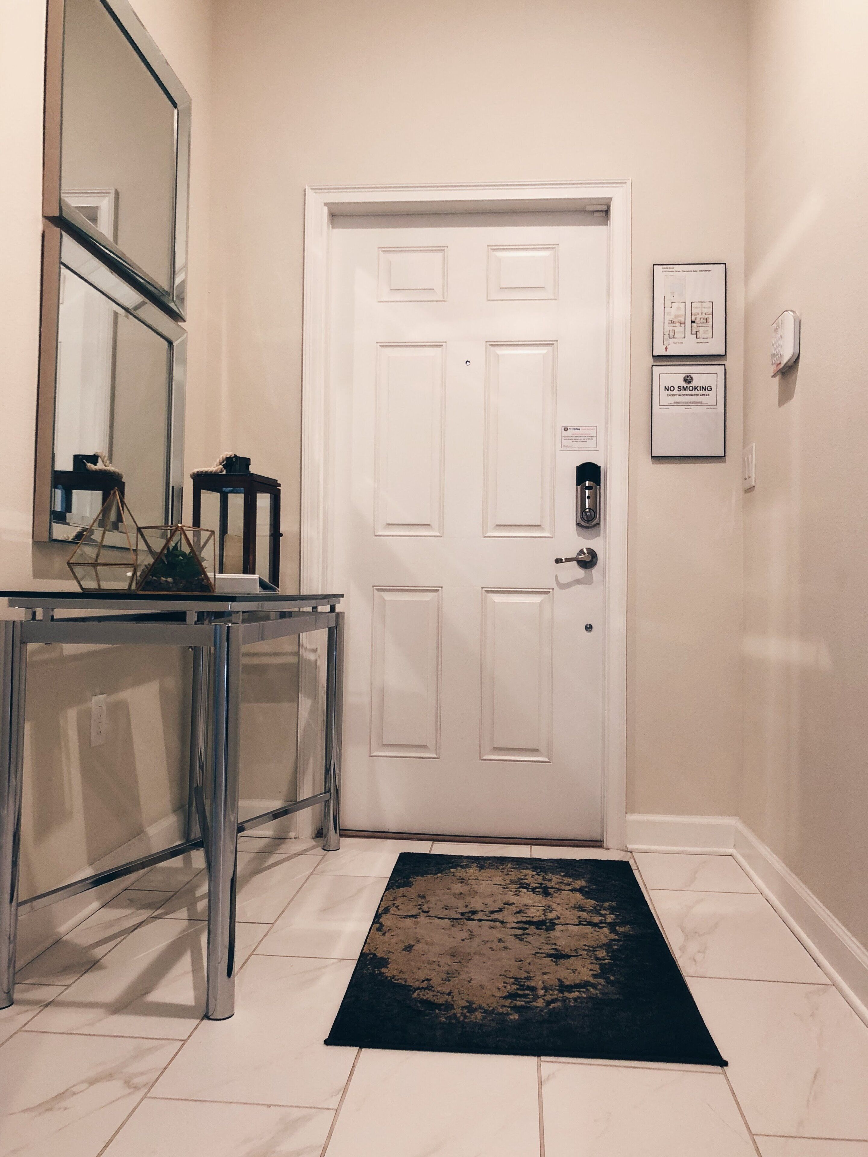 A hallway with a white door, a glass-top console table, a black rug, and a wall-mounted smoke detector.