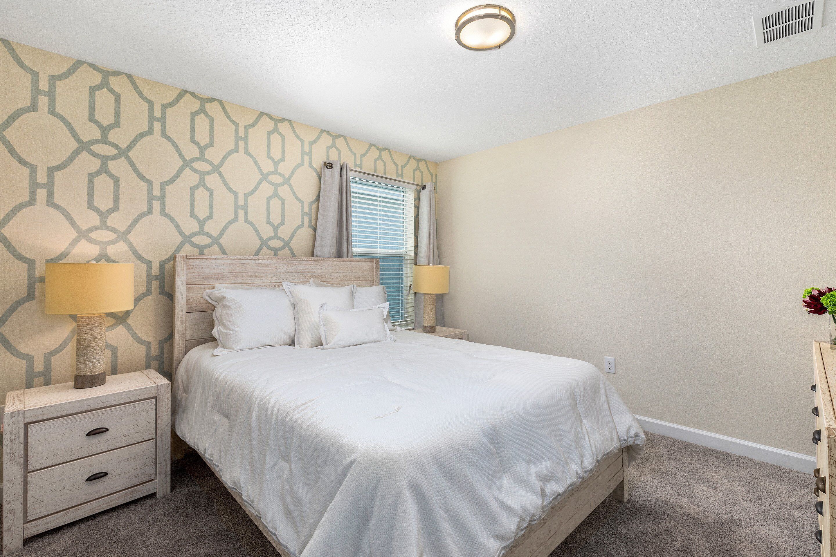A neatly arranged bedroom with a bed, two bedside tables, a nightstand, a window with blinds, and a wall with a geometric pattern.