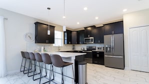 A modern kitchen with dark cabinets, a central island with bar stools, stainless steel appliances, and a trash can. - Last Minute Disney Getaway! Villa with Private Pool & Hot Tub. Book Now! #43253 (Orlando)
