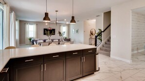 Una cocina moderna con armarios oscuros, una isla grande, iluminación colgante y vistas a una sala de estar con una escalera.