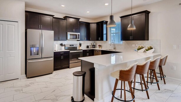 Una cocina moderna con armarios oscuros, electrodomésticos de acero inoxidable, una isla central con taburetes y luces colgantes.