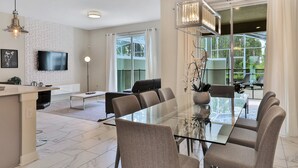 A modern living room with a glass dining table, comfortable seating, a flat-screen TV, and a large window offering a view of the outdoors. - Last Minute Disney Getaway! Private Pool & Themed Bedrooms. Book Now! #43289 (Orlando)