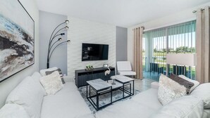 Una sala de estar moderna con un sofá blanco, una mesa de centro, un televisor montado en una pared de ladrillo blanco y un balcón con cortinas.