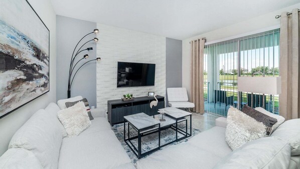 A modern living room with a white sofa, a coffee table, a TV mounted on a white brick wall, and a balcony with curtains. - Last Minute Disney Getaway! Luxury Condo with Private Balcony. Book Now! #44215 (Orlando)