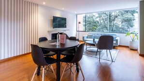 A modern living room with a round dining table, black chairs, a sofa, and a large window with a view of trees. - w* | Homey 2BR in Cabrera (Bogotá)