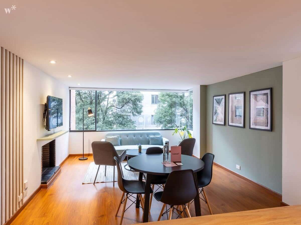 A modern dining area with a round table, black chairs, and a view of trees through large windows.