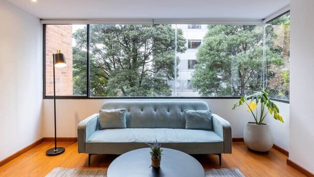 A modern living room with a grey sofa, a round coffee table, and a large window with a view of trees and buildings. - w* | Homey 2BR in Cabrera (Bogotá)