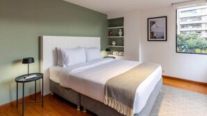 A modern bedroom with a large bed, bedside tables, a nightstand, a window with a view of buildings, and a shelf with decorative items. - w* | Homey 2BR in Cabrera (Bogotá)