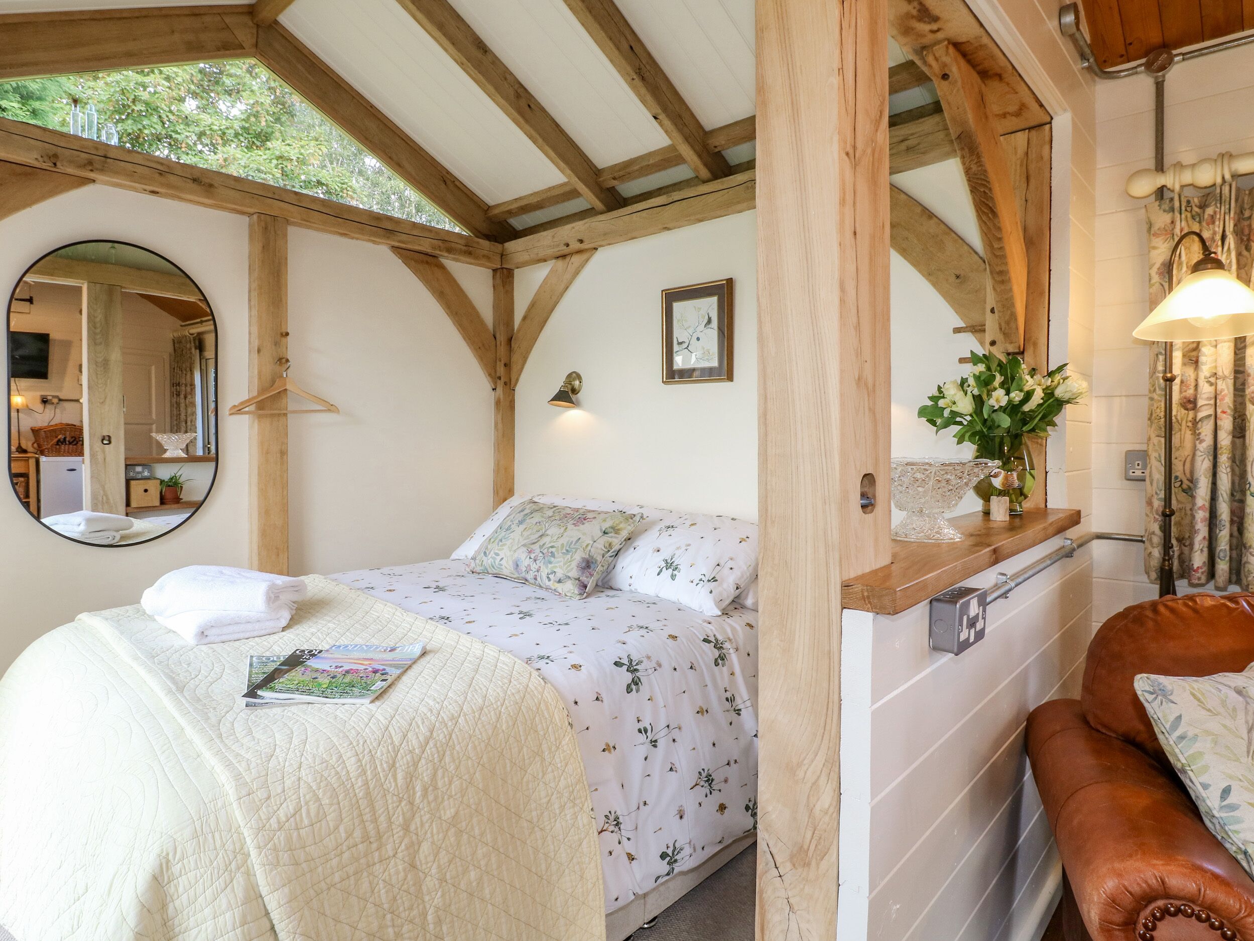 Un dormitorio acogedor con una cama, un espejo, techo de madera y una pequeña mesa con un florero.