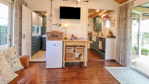 A cozy living room with a wooden ceiling, a white refrigerator, a television, and a dining area with a table and chairs. - The Jenny Wrennery (Rugeley)