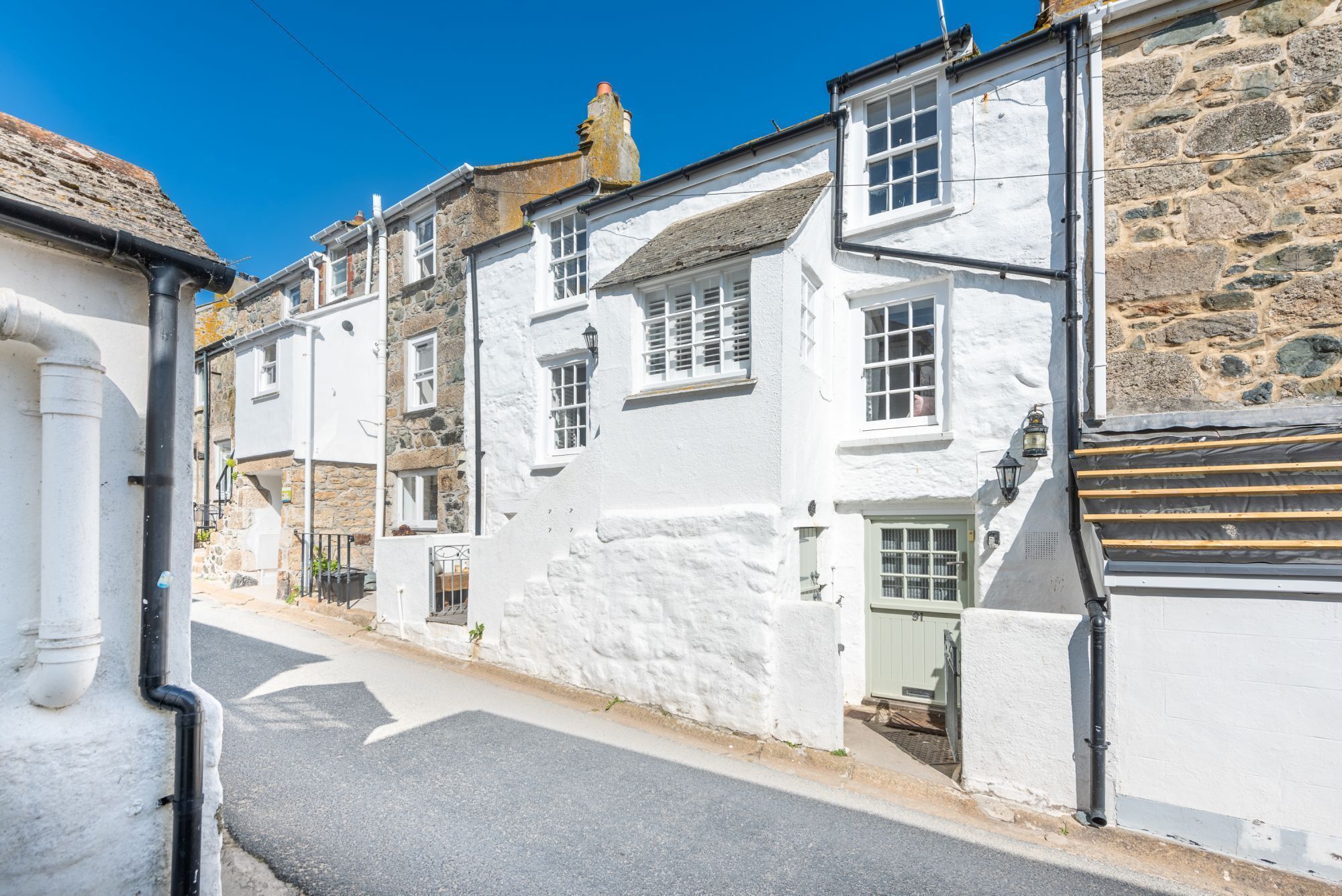 A narrow street lined with white buildings with stone facades, a clear sky, and a paved road.