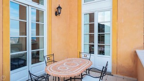 Um pequeno pátio com mesa e cadeiras de azulejos, janelas amplas e um candeeiro fixado na parede.