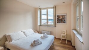 A hotel room with a large bed, a window with a city view, a small white chair, and a framed picture on the wall. - Virgilio 2 - Villa Bice (Alassio)