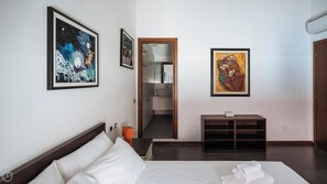 Een hotelkamer met een bed, nachtkastje, klein kastje en een badkamer zichtbaar door een geopende deur.